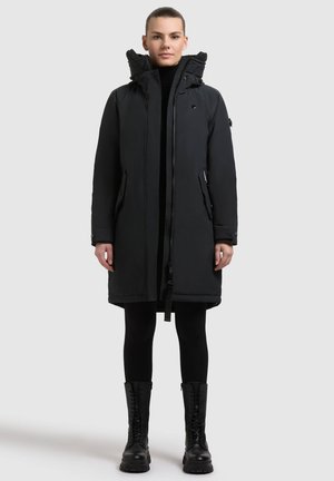 Cappotto parka nero con cappuccio foderato in pelliccia, chiusura con cerniera e due tasche laterali, abbinato a leggins neri e stivali da combattimento con lacci.
