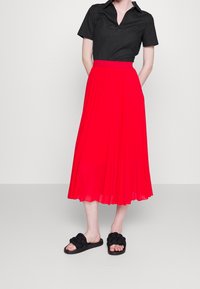 Camicia nera con colletto, gonna midi plissettata rossa e sandali neri con accenti intrecciati. Tessuto liscio, vestibilità strutturata e contrasto di colori vivaci.