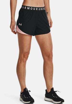Atlet iført sorte og pink Under Armour shorts med logo, kombineret med sorte sportssko, stående mod en hvid baggrund.