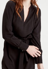 Robe noire à finition texturée, avec un col en V et des manches longues. Comprend une ceinture nouée à la taille et des poignets à boutons.