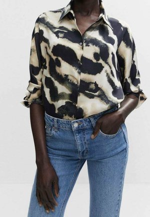 Person wearing a buttoned, black and beige abstract patterned shirt with rolled-up sleeves and high-waisted blue jeans, one hand in pocket.