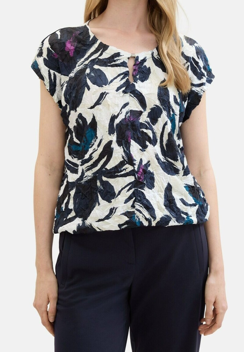 Femme portant un chemisier froissé à manches courtes avec un motif floral abstrait noir, blanc, bleu et violet et un pantalon noir.
