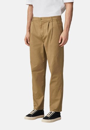 Mężczyzna ubrany w beżowe, plisowane chinosy z bocznymi kieszeniami i czarne trampki, stoi na jednolitym białym tle.