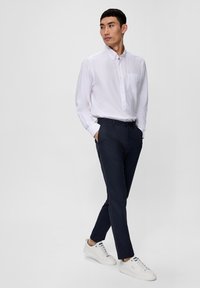 Camicia bianca con bottoni e taschino sul petto, abbinata a pantaloni blu navy e sneakers bianche. Tessuto morbido, vestibilità su misura e linee pulite.