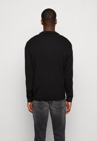 Pull en tricot noir à manches longues et col rond, avec une texture lisse et un design minimaliste, assorti à un jean gris foncé.