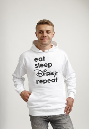 Junger Mann trägt einen weißen Hoodie mit der Aufschrift „eat sleep Disney repeat“, steht vor einem schlichten hellen Hintergrund, Hände in den Taschen.