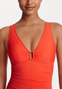 Ljust orange baddräkt i ett stycke med djup V-ringning, samlad front och en dekorativ guldaccent i mitten. Slät yta, justerbara remmar.