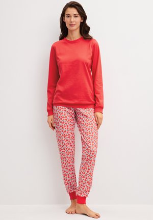 Vrouw draagt een rood shirt met lange mouwen en een witte broek met een rood fruitpatroon, staat blootsvoets tegen een witte achtergrond.