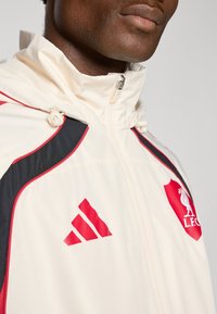 White jacket with red and black accents, zip closure, high collar, featuring adidas and L.F.C. logos on the chest. Smooth fabric texture.