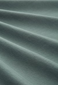 Tekstil med struktur i dæmpet teal farve, der har glatte, foldede lag, der skaber et blødt udseende. Bomulds- eller bomuldsblandingsmateriale.