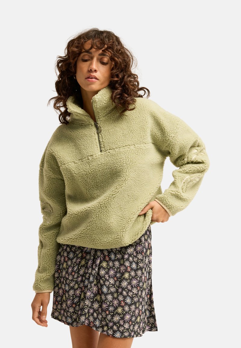 Femme aux cheveux bouclés portant un pull en polaire vert clair et une jupe noire fleurie, debout avec une main sur la hanche.