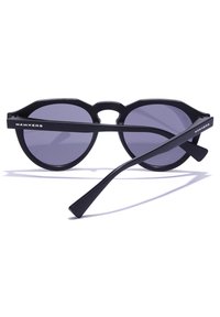 Gafas de sol negras con lentes redondas, que presentan un diseño único de montura octagonal y varillas negras delgadas. El nombre de la marca "Hawkers" es visible en los lados.