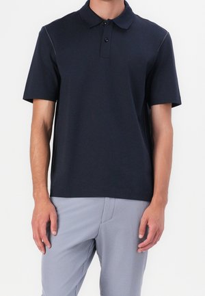 Man wearing a plain dark navy short-sleeve polo shirt and light gray pants, standing with arms relaxed by sides.