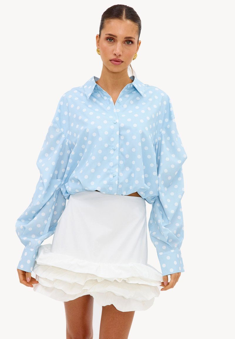 Femme portant une chemise oversize bleu clair à pois blancs et une jupe mini blanche à ourlet volanté superposé, debout sur un fond uni.