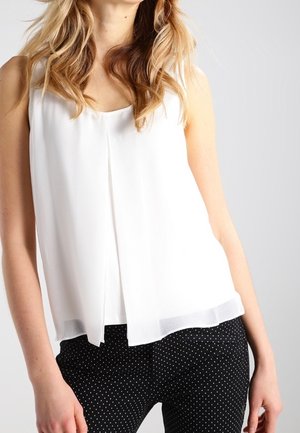 Blouse blanche sans manches avec un ourlet droit et un tombé souple ; portée avec un pantalon noir à pois blancs petits.