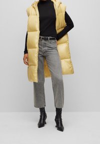 Veste sans manches en polaire jaune avec capuche, haut à manches longues noir, jean gris à jambes larges et bottines noires. Texture lisse et design minimal.