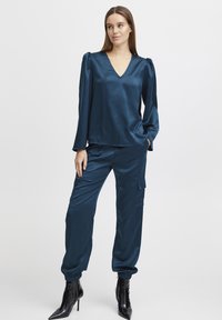 Bluză din satin turquoise cu decolteu în V și mâneci bombate, asortată cu pantaloni cargo tapered. Pantalonii au manșete elastice și buzunare.