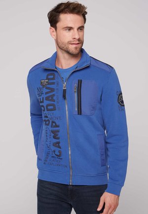 Maglione blu con zip e colletto alto, dotato di una tasca sul petto, con accenti testurizzati e dettagli stampati sulla parte anteriore.