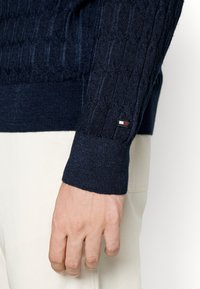 Gros plan sur une main portant une manche de pull torsadé bleu marine avec un petit patch logo rectangulaire au-dessus du poignet, associée à un pantalon blanc.