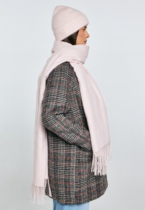 Žena v profilu, oblečená v kostkovaný kabát, světle růžovou pletenou čepici a odpovídající dlouhou šálu na jednobarevném světlém pozadí.