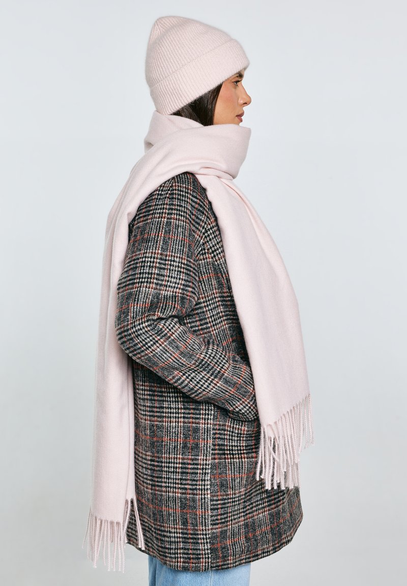 Profiilifoto naisest, kes kannab ruudulist mantlit, kahvaturoosat kootud mütsi ja sobiva pikkade salliga, taustal hele ühevärviline taust.