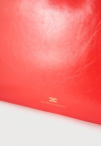 Κόκκινη υφή δερμάτινης επιφάνειας με μικρό χρυσό λογότυπο «Elisabetta Franchi» και αρχικά κοντά στην κάτω άκρη, σε λευκό φόντο.