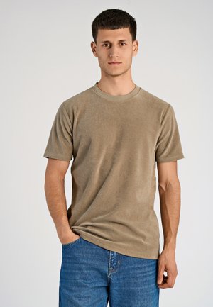 Beżowa koszulka z okrągłym dekoltem z miękkiego materiału, z krótkimi rękawami, o klasycznym kroju. Model zestawia ją z niebieskimi dżinsami.