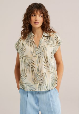 Kobieta o kręconych włosach, ubrana w koszulę z krótkim rękawem zapinaną na guziki z zielonym motywem liści oraz jasnoniebieskie spodnie, z rękami w kieszeniach, patrząca w bok.