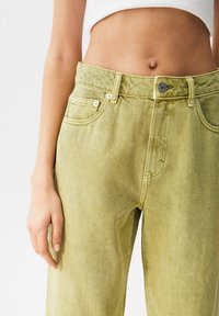 Jean en denim vert clair avec une taille haute, doté de poches avant, de ferrures métalliques et d'une finition texturée.
