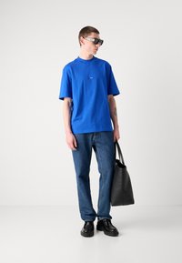 Blå bomull T-skjorte med rund hals, ledsaget av denimjeans og svarte lærsko. En svart totebag holdes i venstre hånd.