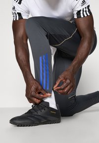 Sorte atletiske sko med teksturerede såler, grå atletiske leggings med blå striber, og en hvid atletisk skjorte med sorte detaljer.