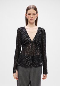 Γυναίκα με ίσια καστανά μαλλιά, που φορά διαφανή μαύρη μπλούζα με σχέδιο και μακριά μανίκια, με βαθύ V-λαιμό και γκρι ραμμένο παντελόνι.