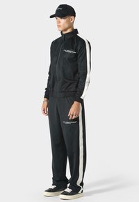 Czarny dres z miękkiego materiału z białymi paskami po bokach, kurtka na zamek i elastyczny pas. Zawiera detale z logo oraz czarną czapkę.