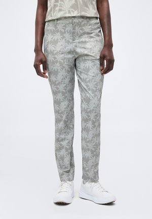 Person iført lysegrå bukser med et diskret palmetræsmønster, en matchende skjorte og hvide sneakers på en hvid baggrund.