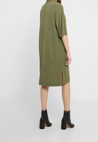 Robe midi verte olive avec des manches courtes, détail d'ourlet fendu et texture douce. Associée à des bottines à talons noires.