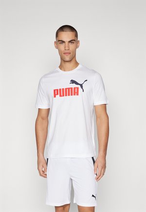 Giovane uomo che indossa una maglietta bianca Puma con logo rosso e pantaloncini bianchi Puma, in piedi davanti a uno sfondo semplice.