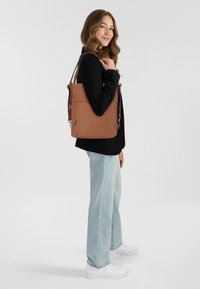 Bolso de hombro de cuero marrón con correas ajustables, acabado texturizado y un bolsillo con cremallera. Lleva puesto con pantalones vaqueros de color azul claro y zapatillas blancas.