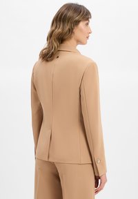 Femme aux cheveux ondulés portant un costume beige ajusté avec des détails de boutons au dos et aux poignets, se tenant devant un fond blanc.