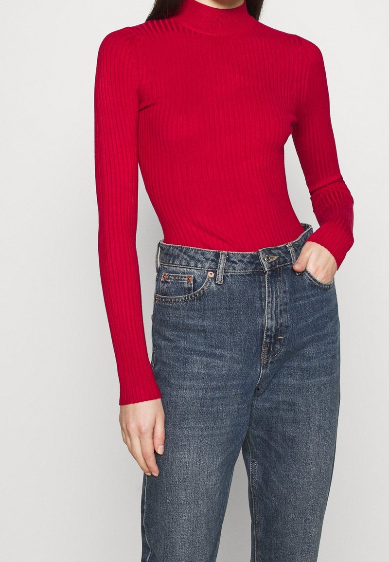 Femme portant un pull à col roulé côtelé rouge ajusté et un jean bleu taille haute, une main dans la poche, sur un fond uni.