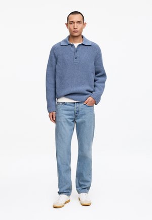 Mann steht und trägt einen blauen Strickpullover mit Kragen, hellblaue Jeans, weiße Sneaker und ein darunter hervorschauendes weißes Hemd.