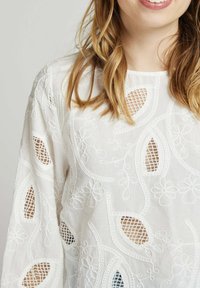 Blouse blanche brodée avec des motifs floraux et de feuilles, dotée de découpes. Fabriquée en tissu léger avec une légère texture.