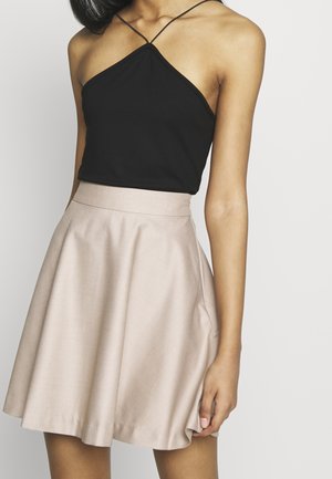 Mujer con top sin mangas negro de estilo halter con tirantes delgados y una falda beige de línea A de talle alto, de pie contra un fondo liso.