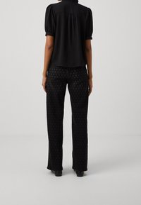 Blusa negra de mangas abullonadas con la parte trasera ligeramente fruncida, combinada con jeans negros que presentan un sutil patrón de puntos y un diseño de pierna recta.