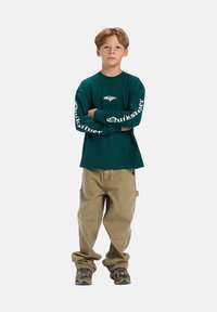 Teal T-shirt met lange mouwen met wit "Quiksilver" logo op de mouwen, gecombineerd met lichtbeige, losse pasvorm broek en schoenen met camouflagepatroon.