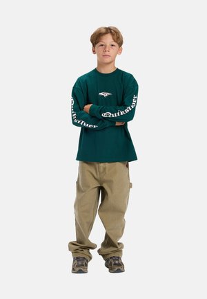 Teal T-shirt met lange mouwen met wit "Quiksilver" logo op de mouwen, gecombineerd met lichtbeige, losse pasvorm broek en schoenen met camouflagepatroon.