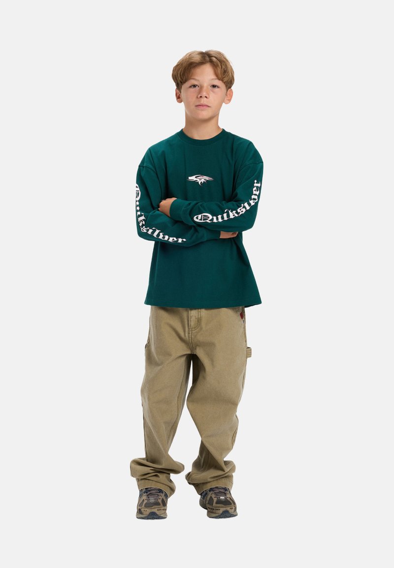 Teal T-shirt met lange mouwen met wit "Quiksilver" logo op de mouwen, gecombineerd met lichtbeige, losse pasvorm broek en schoenen met camouflagepatroon.