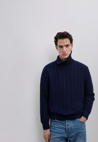 Pull en col roulé en maille torsadée navy avec un motif texturé et un ourlet côtelé, porté avec un jean bleu clair, sur un fond uni.