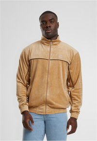 Bežna žametna jakna na zadrgo z visokim ovratnikom, črnimi akcentnimi črtami in elastičnimi manšetami. Diskretna logotip na zgornji levi strani. Gladka tekstura.