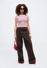 Γυναίκα με σγουρά μαλλιά που φοράει γυαλιά ηλίου, κόκκινη και λευκή ριγέ κοντή μπλούζα, φαρδιά καφέ παντελόνια, κόκκινη τσάντα και κόκκινα παπούτσια, στέκεται κοιτώντας μπροστά.