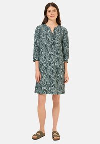 Vihreä mekko, jossa on lehdenkuvio, 3/4-hihat ja kauluskanta. Kangas näyttää kevyeltä ja pehmeältä. Yhdistetty ruskeisiin sandaaleihin.
